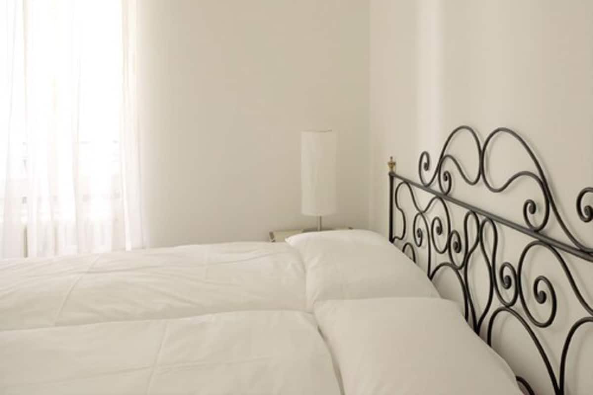 Un lit avec une tête de lit en métal, dans une pièce dont un mur est peint en blanc et qui possède une fenêtre ornée de rideaux légers.