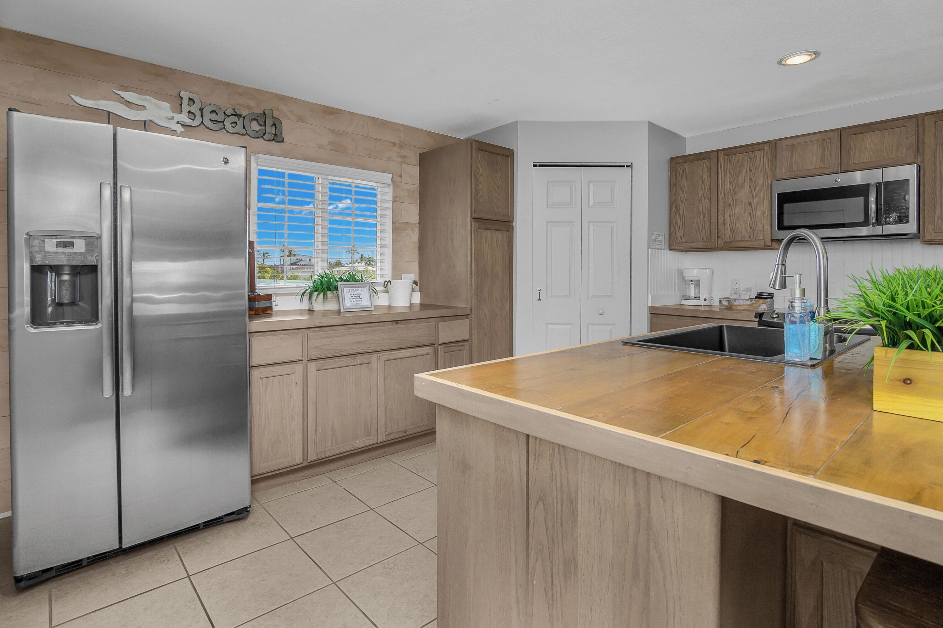 Una cocina con refrigerador de acero inoxidable, gabinetes de madera, microondas y una ventana con persianas.