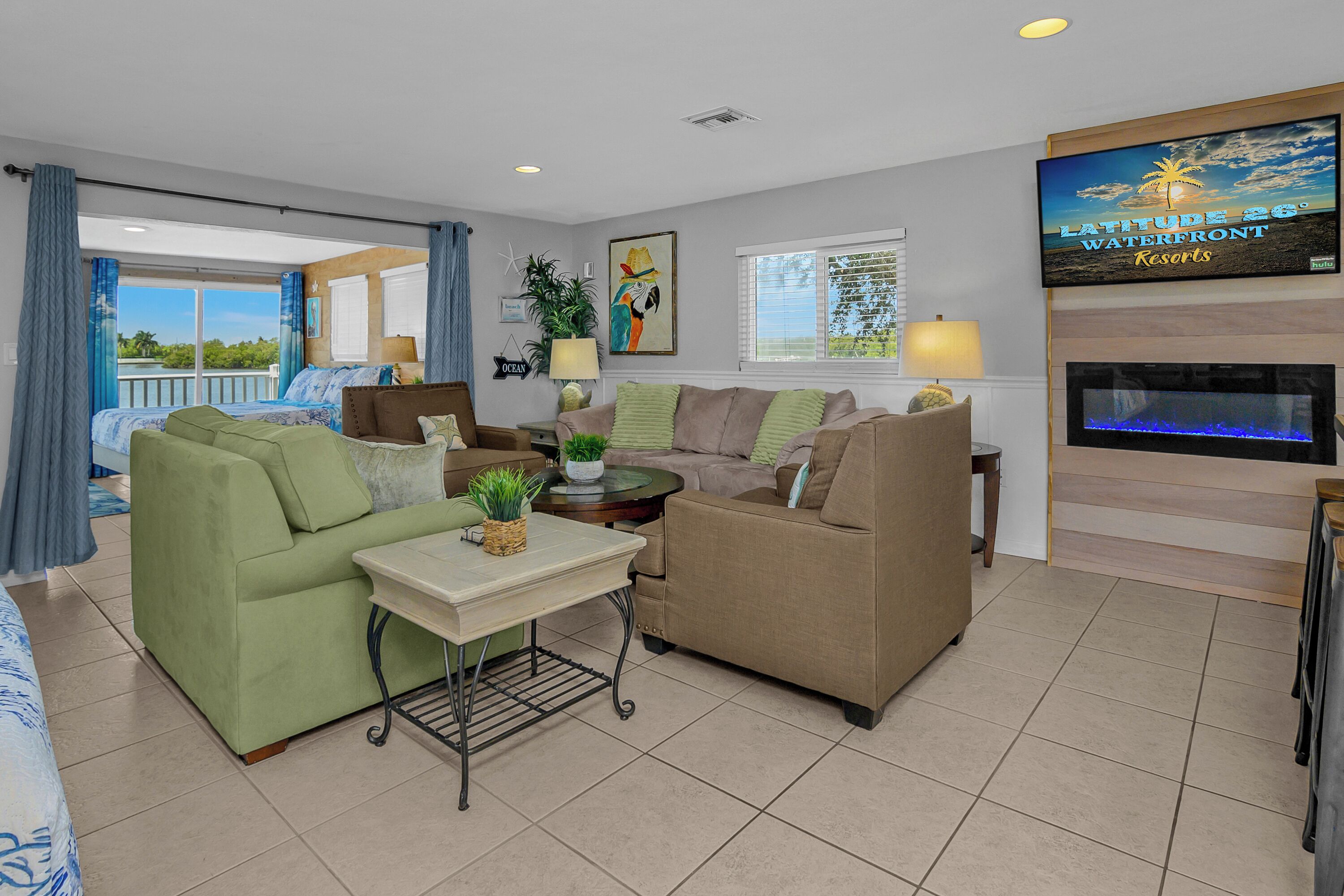 A living room with a sofa, armchair, coffee table, and TV. There's a fireplace, a balcony with curtains, and a painting on the wall.