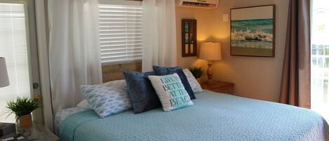 A bedroom with a bed, a nightstand, a lamp, a window with blinds, a wall-mounted air conditioner, and a beach-themed pillow.