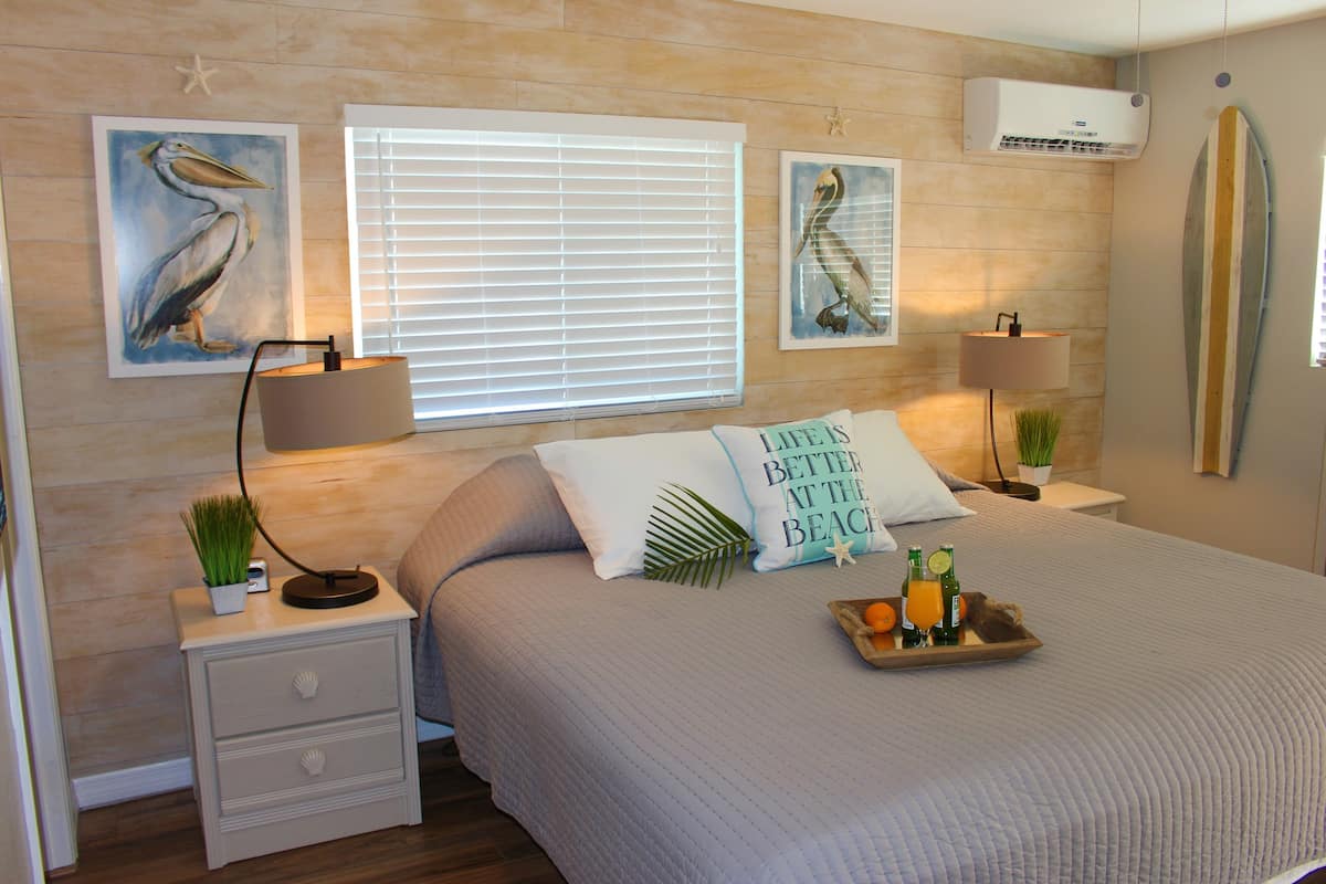 Een slaapkamer met een bed, twee nachtkastjes, een raam met zonwering, wanddecoratie, een airconditioner en een surfplank.