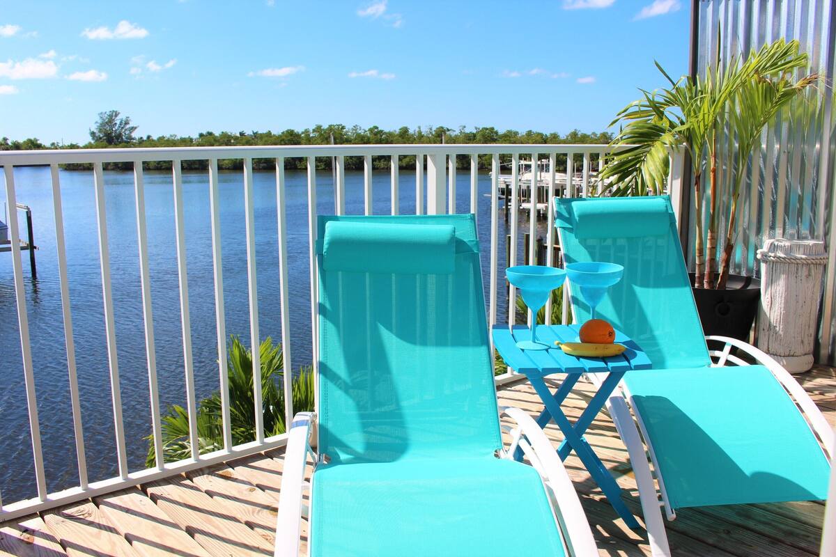 Een terras met twee turquoise ligstoelen, een tafel met een banaan en een sinaasappel, en uitzicht op het water.