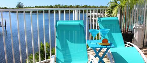 A deck with two turquoise lounge chairs, a small table with a banana and an orange, and a view of the water and dock.