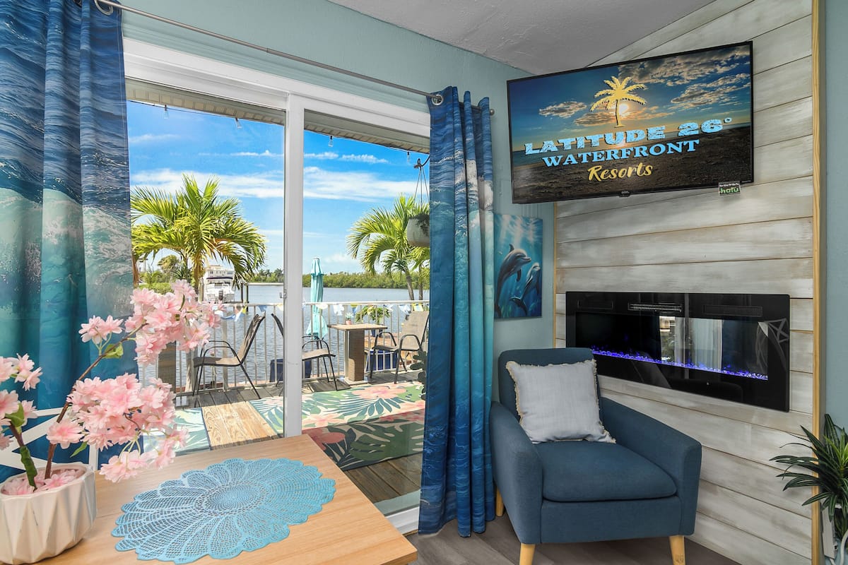 Een kamer met een televisie waarop 'Latitude 26° Waterfront Resorts' staat, een blauwe bank, een houten bijzettafeltje, een decoratieve kantborduurwerk en uitzicht op het landschap met palmbomen en een heldere lucht.