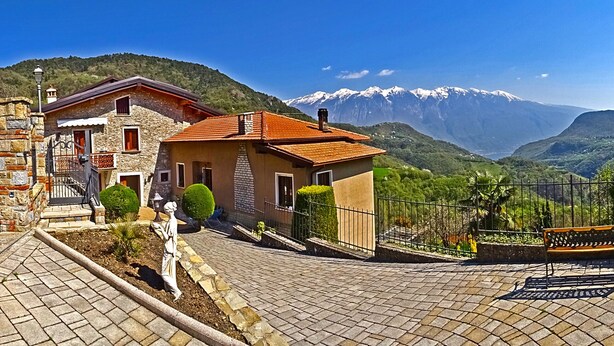 A stone house with a red-tiled roof, a paved courtyard, and a mountain backdrop.