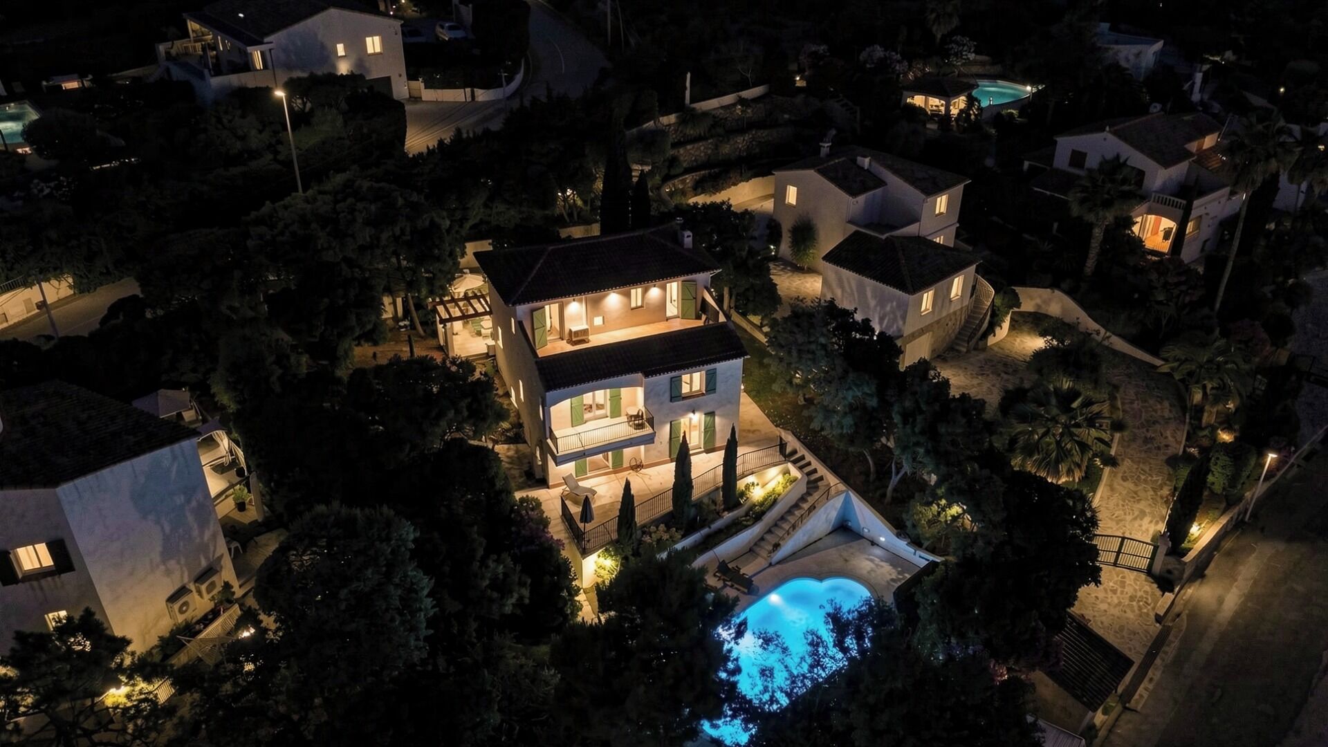 A large, multi-story house with a swimming pool, surrounded by trees and other houses.