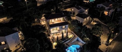 Uma casa grande, com vários andares, piscina e cercada por árvores e outras casas.