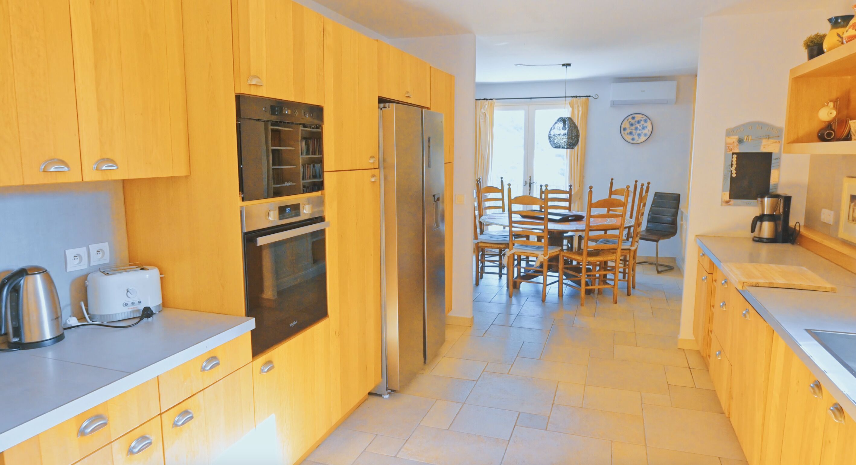A kitchen with wooden cabinets, a refrigerator, an oven, a dining table with chairs, and a coffee machine.