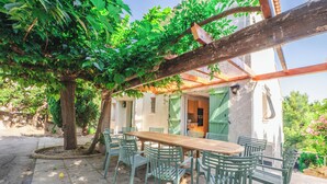 Un espace de restauration en plein air avec une table en bois et des chaises vertes, entouré d’une pergola et d’arbres.
