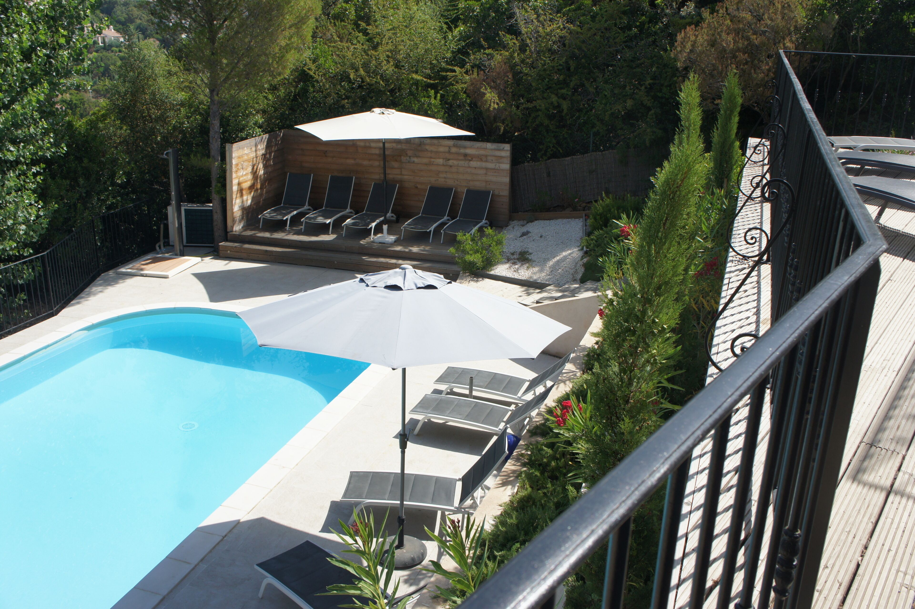 Piscina con sillones de descanso y sombrillas, rodeada de una terraza de madera y vegetación.