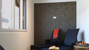 A room with a brick-patterned wall, a window with blinds, and two wicker chairs with a red towel on one. - Lovely apartment for 4 people with WIFI, hot tub, TV and balcony (Leukerbad)