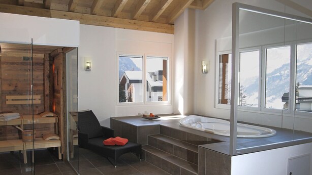 A modern bathroom with a wooden ceiling, a glass shower enclosure, a black chair, a large mirror, and a view of mountains through the windows. - Lovely apartment for 4 people with WIFI, hot tub, TV and balcony (Leukerbad)
