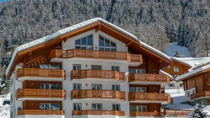 A multi-story hotel with wooden balconies and large windows, surrounded by snow-covered trees and mountains. - Lovely apartment for 4 people with WIFI, hot tub, TV and balcony (Leukerbad)
