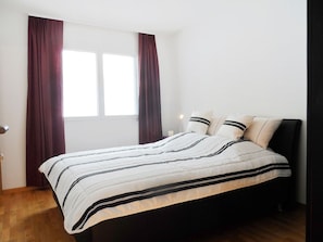 Ein Schlafzimmer mit einem Bett, zwei Fenstern und einem Nachttisch mit Lampe.