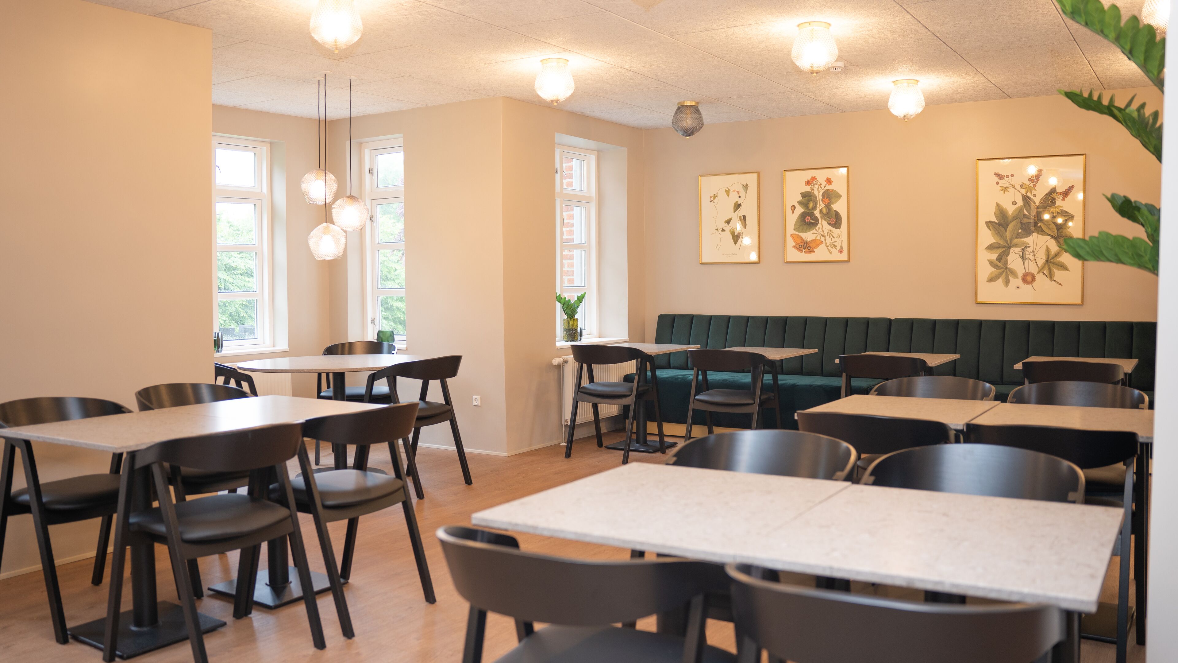 A restaurant with multiple tables and chairs, a green booth seating area, and framed botanical artwork on the walls.