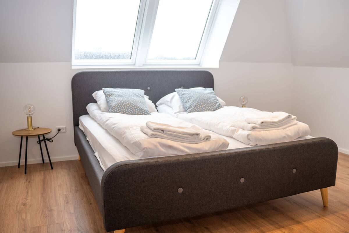 A neatly made bed with white linens and two pillows, a small round table with a lamp, and a window above the bed.