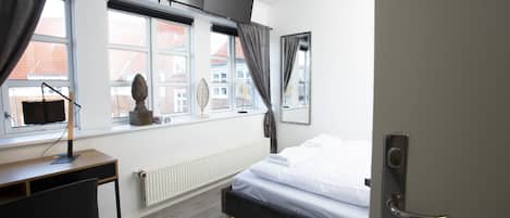 A bedroom with a bed, a desk, a computer, and a window with curtains.