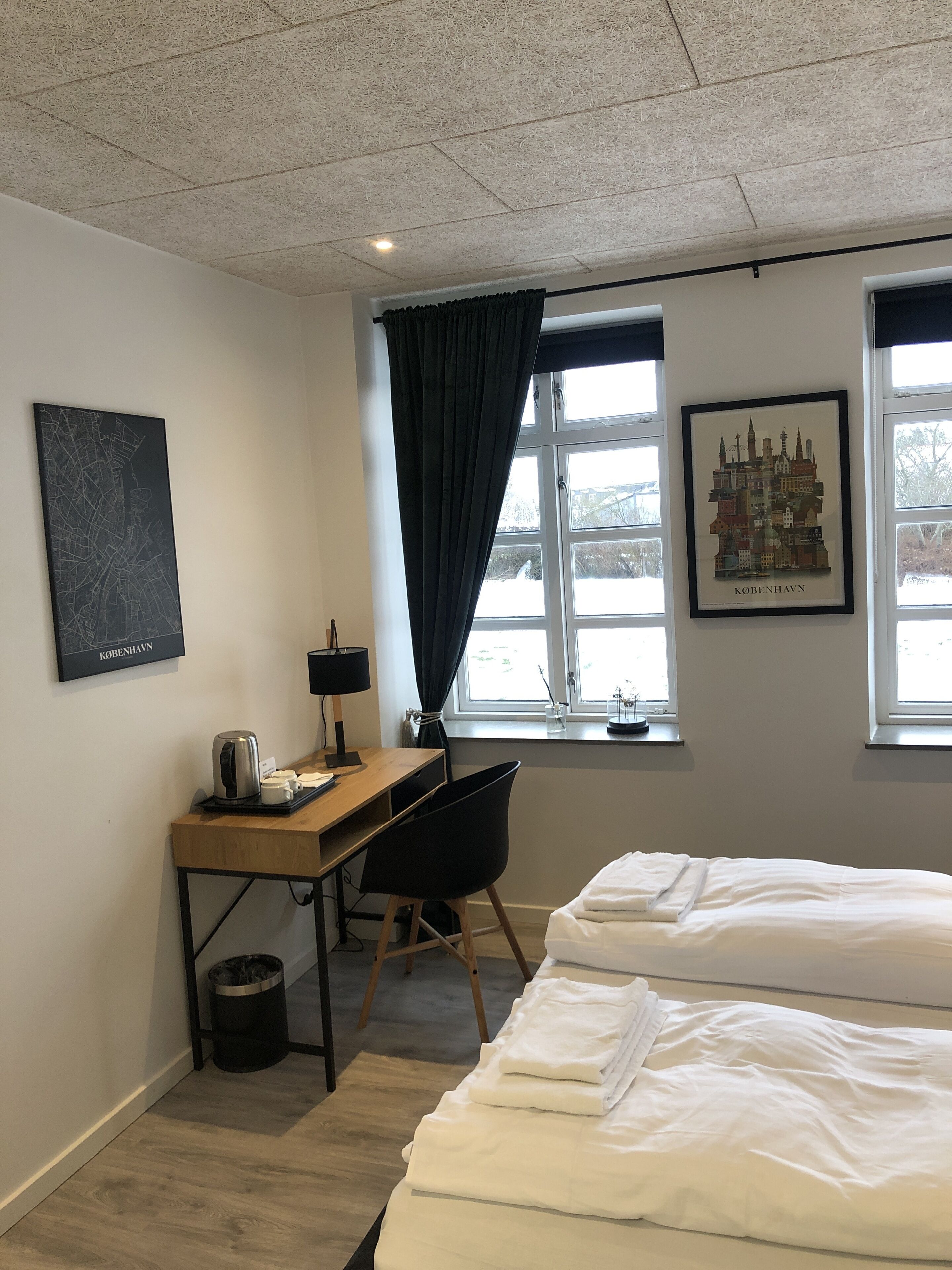 Una habitación de hotel con una cama, un escritorio con una silla, una cafetera y un cuadro enmarcado con una vista urbana.