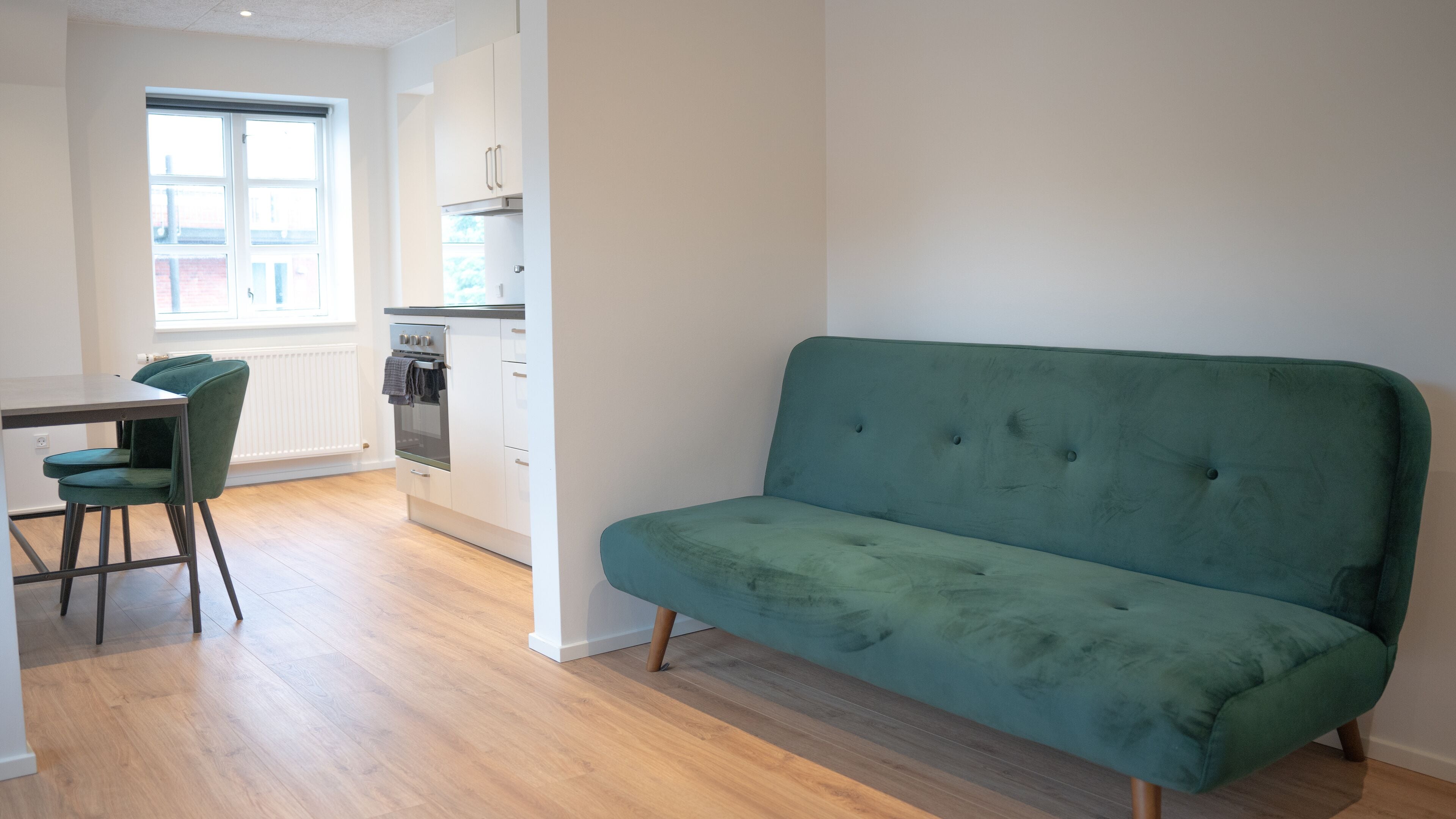 A modern kitchen with white cabinets, a built-in oven, and a window. There is a green sofa and two green chairs in a living area.