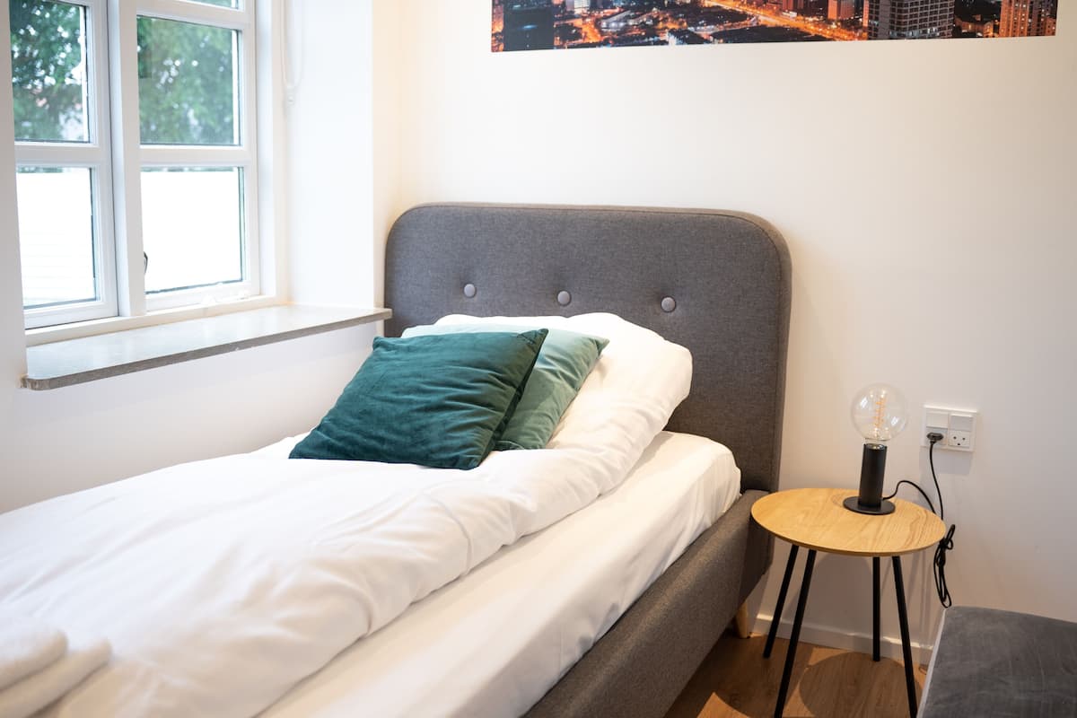 A neatly made bed with white and teal pillows, a grey upholstered headboard, and a small round wooden table with a lamp beside it.