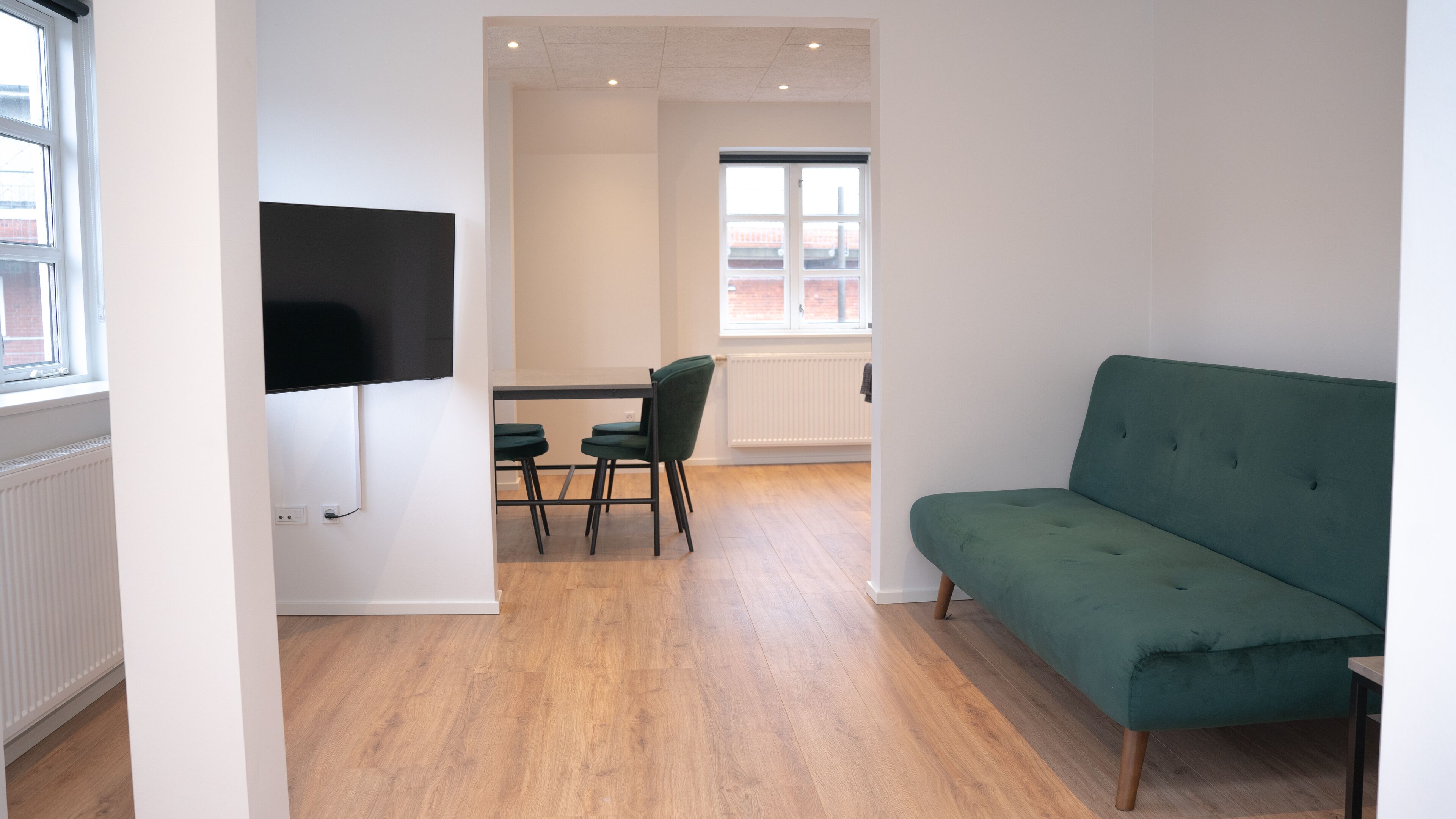 A modern living room with a green sofa, a flat-screen TV mounted on the wall, a dining area with a table and chairs, and a window with a view of a brick building.