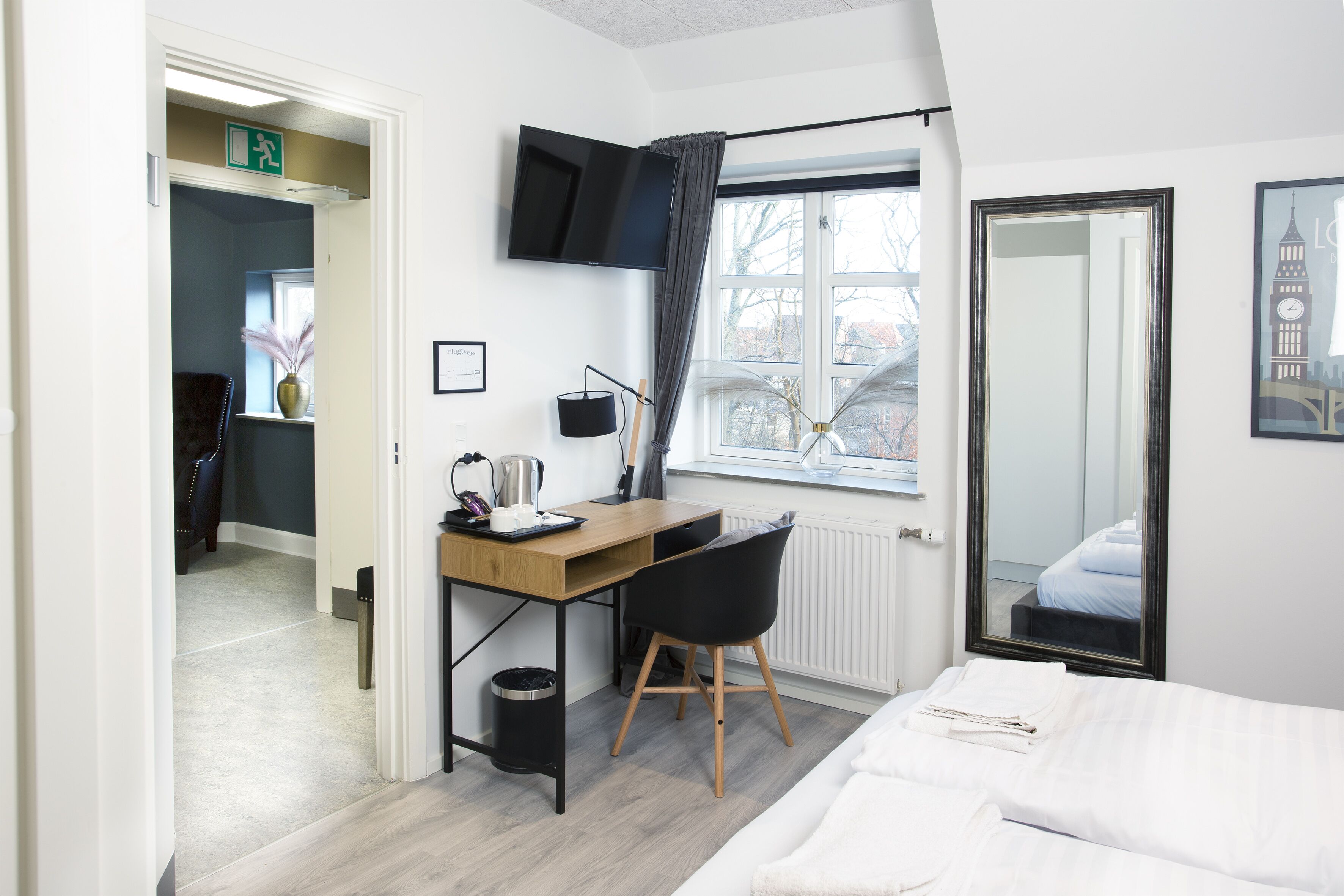Quarto de hotel moderno com escrivaninha de madeira, cadeira preta e cama com roupa de cama branca.