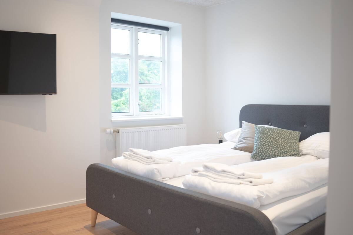 A bedroom with a bed, a window, a radiator, and a flat-screen TV mounted on the wall.
