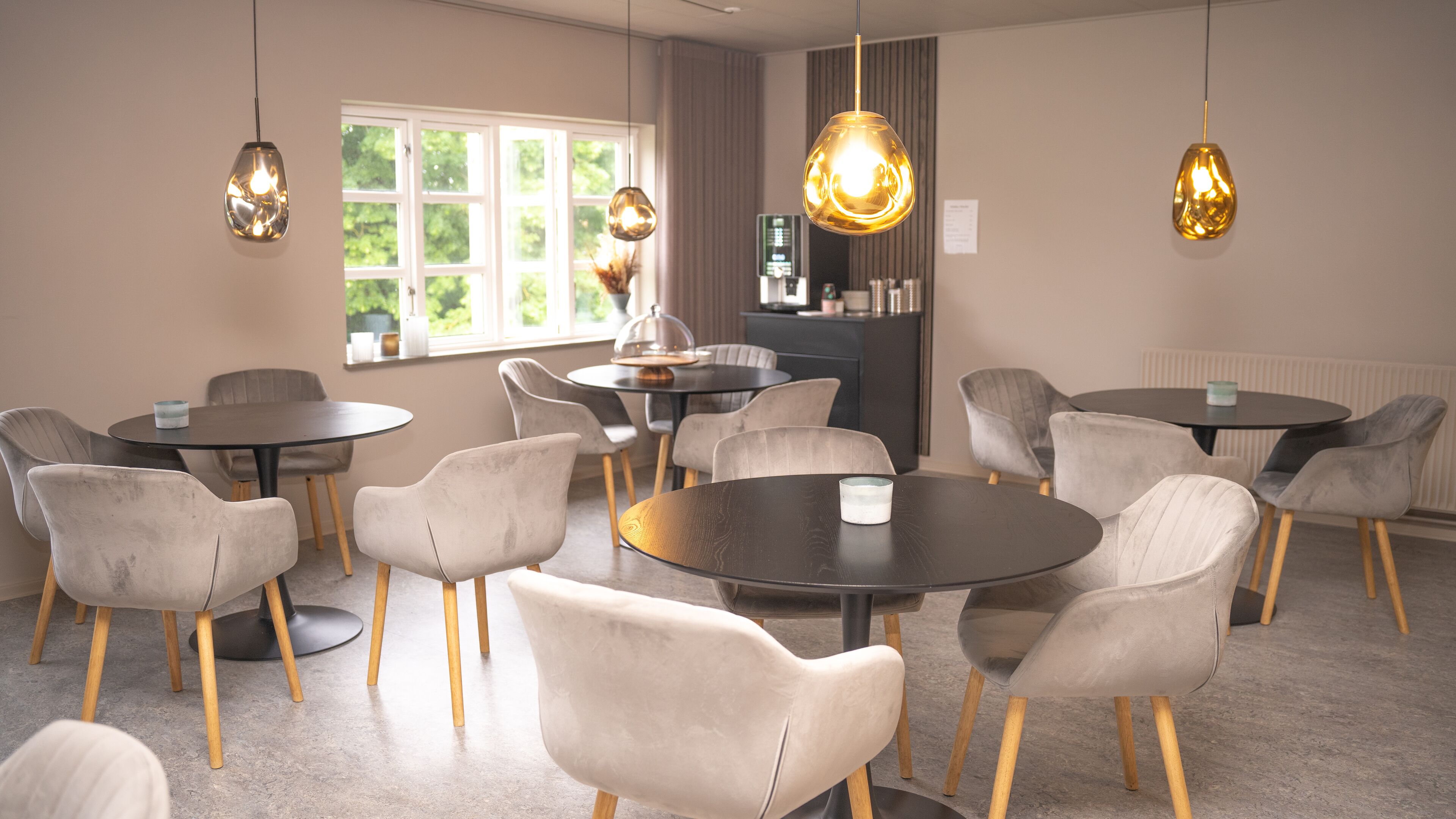 A modern dining area with round tables and cushioned chairs, pendant lighting, and a view of greenery through large windows.
