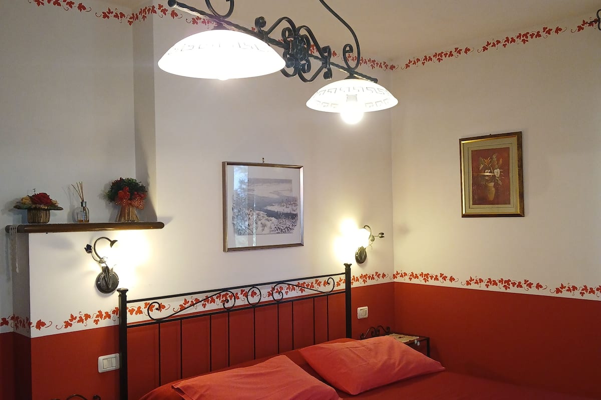 Une chambre à coucher avec une couvre-lit rouge, une table de chevet, une étagère murale ornée d’objets décoratifs et un luminaire fixé au plafond.