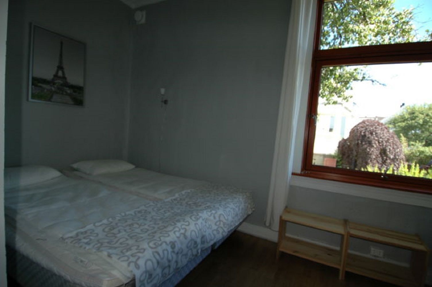Een slaapkamer met een bed, een raam met uitzicht op groen, en een klein houten bankje.