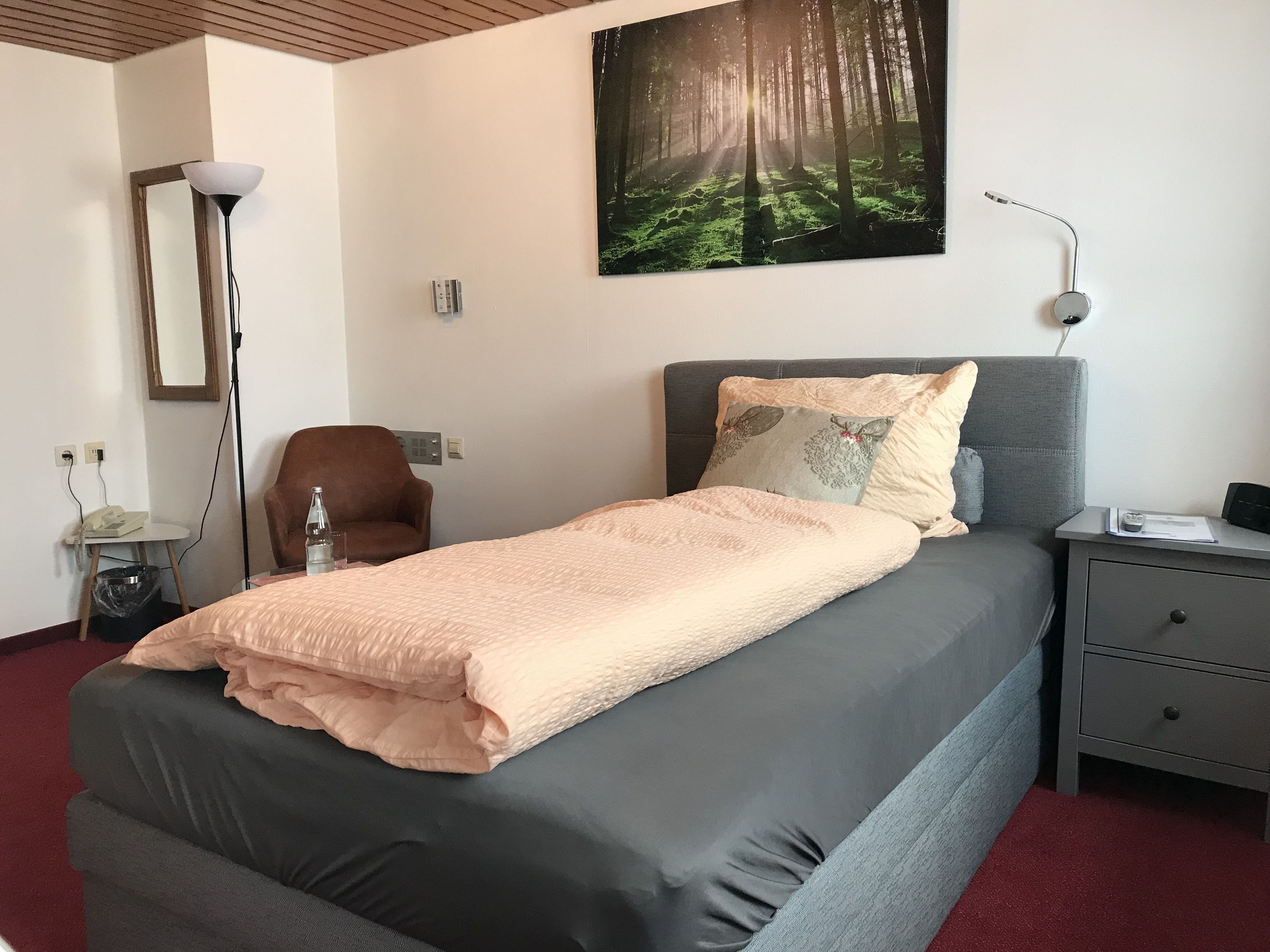 A hotel room with a bed, a chair, a desk, and a painting of a forest.