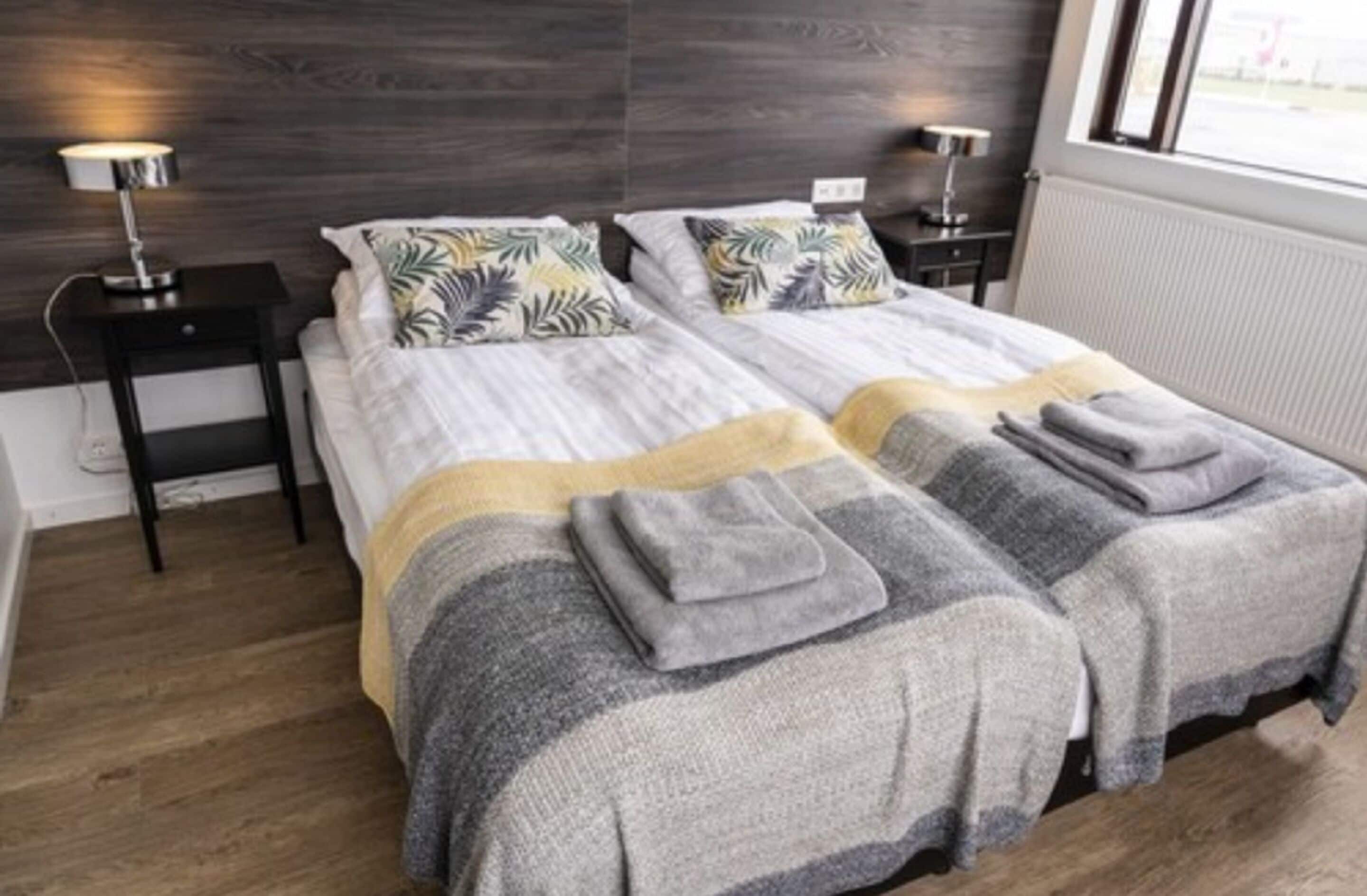 A double bed with a striped blanket and two pillows, a bedside table with a lamp, and a window with a view of the outside.