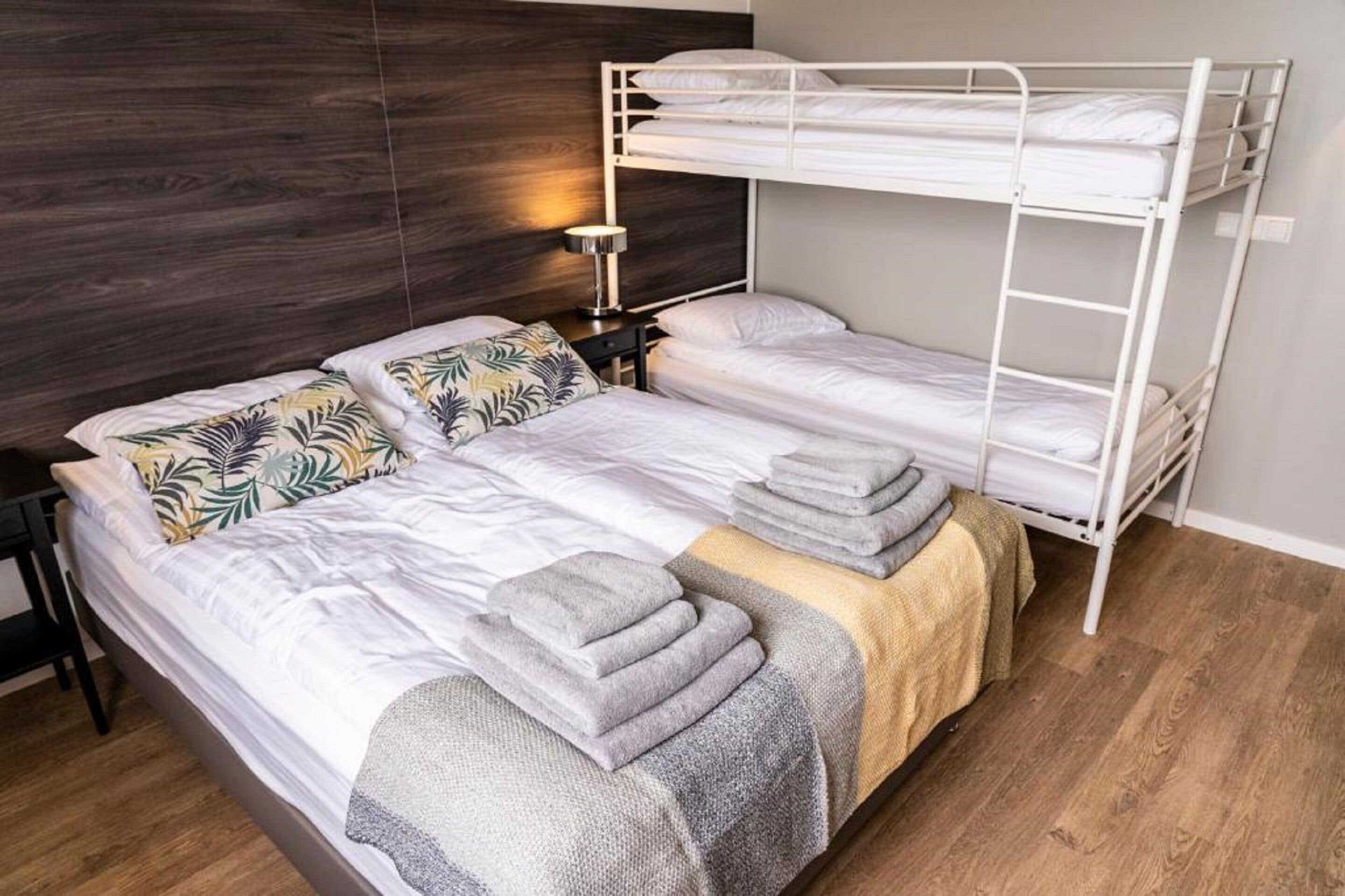 Habitación con literas, cabecera de madera, mesita de noche con lámpara y toallas dobladas cuidadosamente.