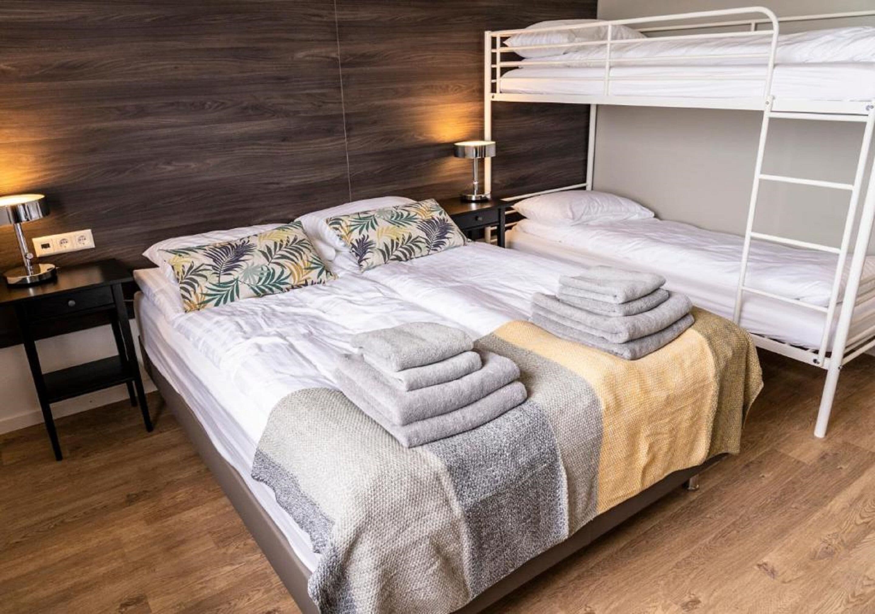 A bedroom with a bunk bed, a bedside table with a lamp, and a bed with a patterned pillow and folded towels.