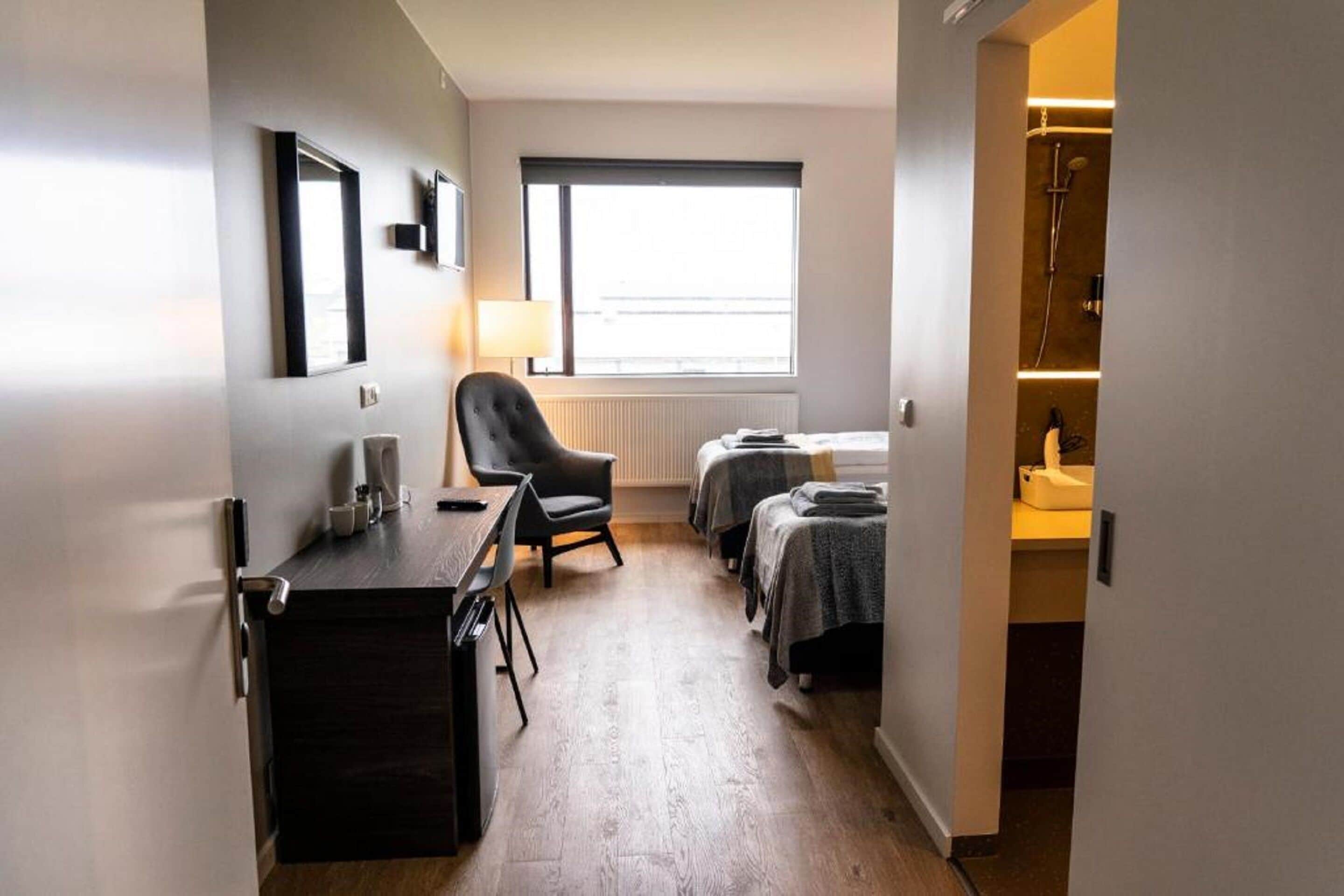 Habitación de hotel con escritorio, silla, dos camas, ventana y baño con ducha.