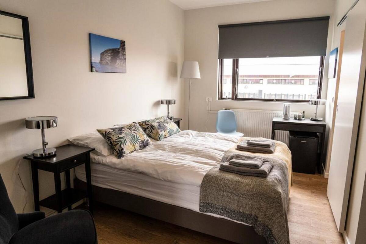 A hotel room with a bed, bedside table, lamp, and a window with a view of buildings.