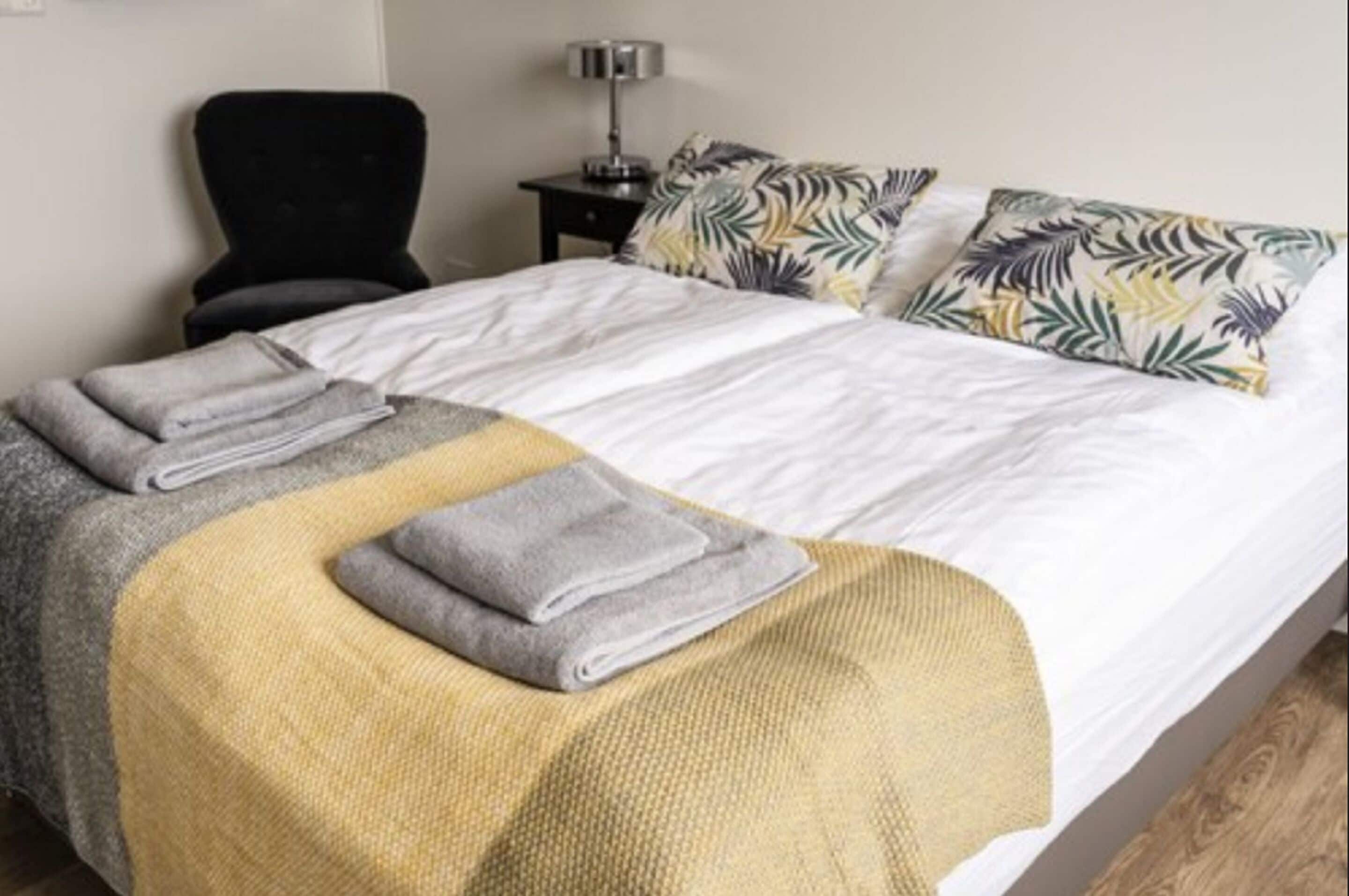 A neatly made bed with a white and yellow blanket, two pillows with a tropical leaf pattern, and a black chair beside the bed.