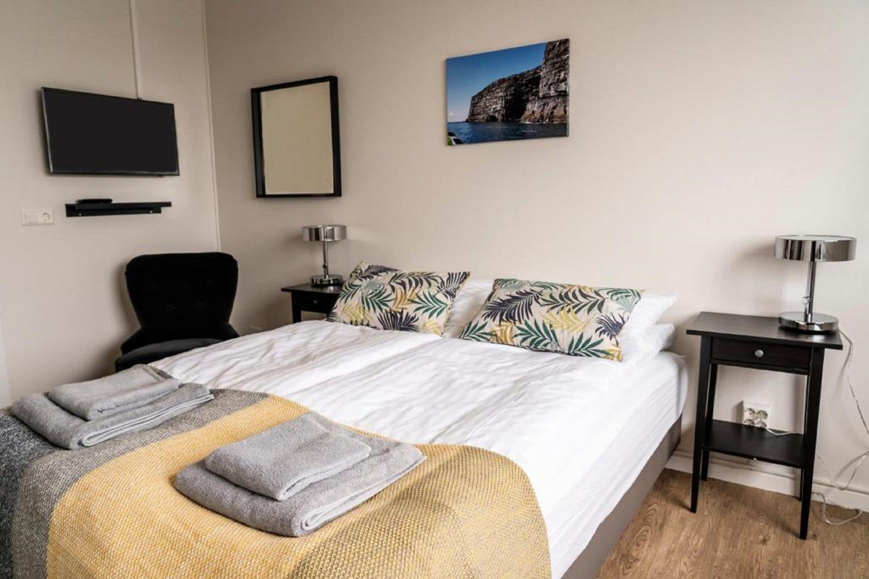 A neatly made bed with a white duvet, pillows with tropical leaf patterns, and folded towels. A black chair is against the wall, and a small table with a lamp is beside the bed.