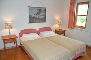 Uma cama de casal com colcha bege e branca, duas mesas de cabeceira com luminárias, um quadro na parede e uma janela com cortina.