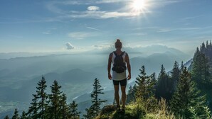 Une personne se tient au sommet d’une montagne, un sac à dos sur le dos, et contemple une vallée parsemée d’arbres et bordée de montagnes lointaines.