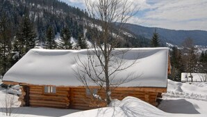 A log cabin with a snow-covered roof, surrounded by a snowy landscape and a forested mountain range.