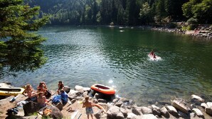 Escena junto al lago con gente sentada sobre rocas, una persona nadando y un bote en el agua.