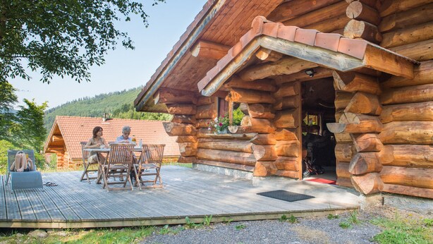 Una cabaña de troncos con terraza de madera, mobiliario exterior y montañas al fondo.