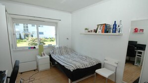 A bedroom with a bed, a desk, a chair, a window, and a shelf with books and bottles.