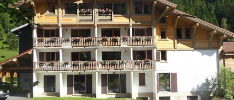 A multi-story wooden building with balconies, likely a hotel or guesthouse, surrounded by greenery and a mountainous backdrop.