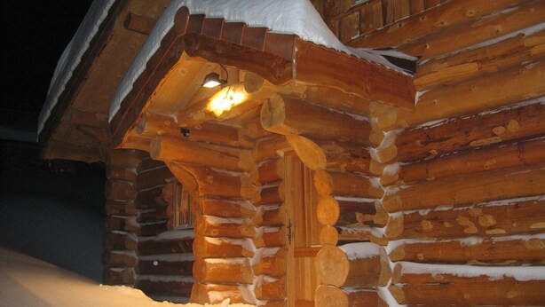 Une cabane en rondins dont l’entrée est recouverte de neige et une fenêtre illuminée.