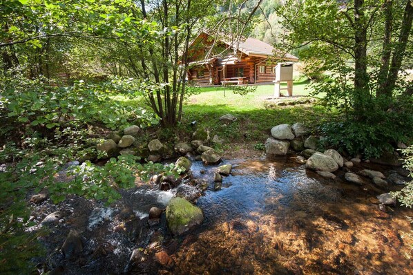 Een rustieke hut omgeven door een weelderig bos, een beek met rotsen en een grasveld.
