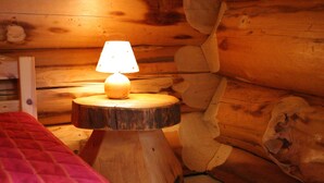 Une chambre confortable dans un chalet, avec un lit en bois, une petite table en bois surmontée d’une lampe et un mur fait de rondins de bois.