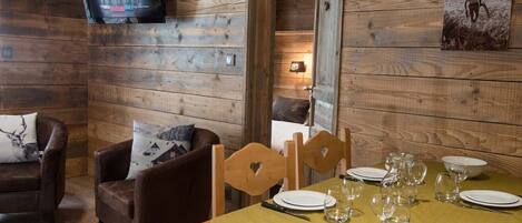 A wooden cabin interior with a dining table set for four, a television mounted on the wall, and a framed picture on the wall.