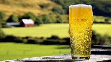 Een glas bier met een etiket "Hawkstone" op een houten ondergrond, met een landelijk landschap op de achtergrond.